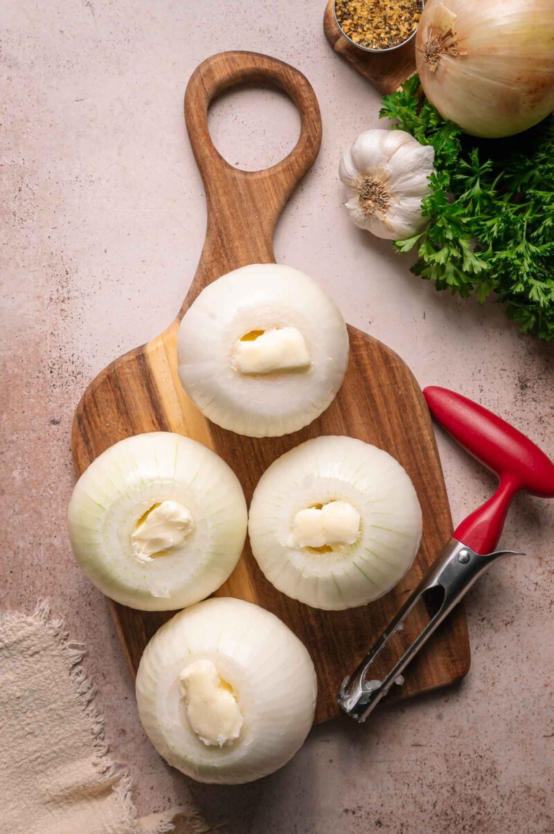 Four whole onions with centers removed and filled with butter are arranged on a wooden cutting board. Nearby are parsley, garlic, an onion, spices, and a vegetable peeler.