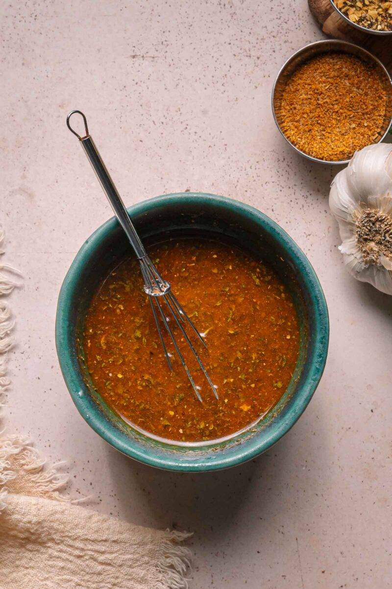 A whisk sits in a green bowl filled with seasoned butter sauce; garlic and a bowl of spices are nearby on a light surface.