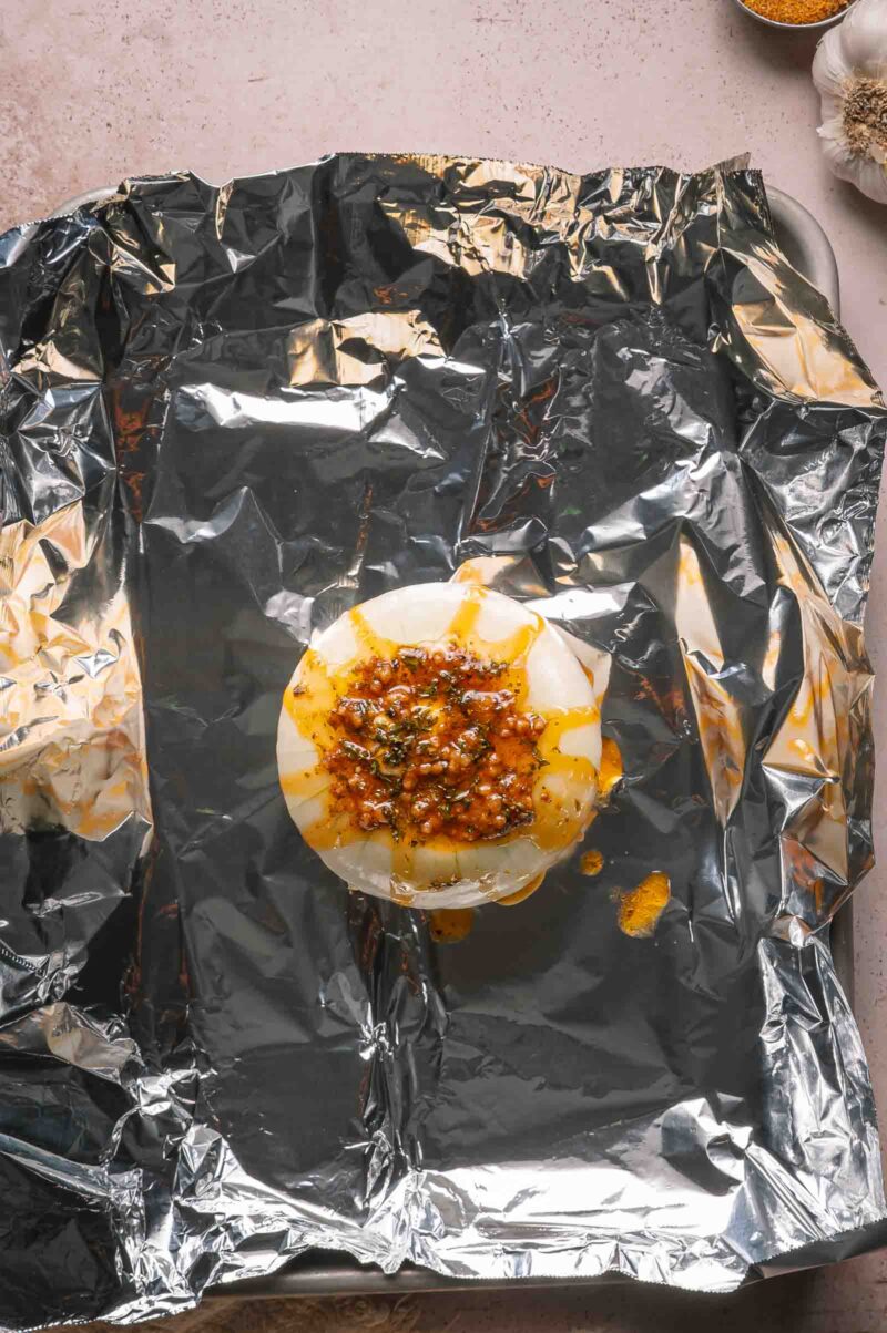A whole onion topped with seasoning and butter sits on a sheet of aluminum foil, ready for wrapping and cooking.