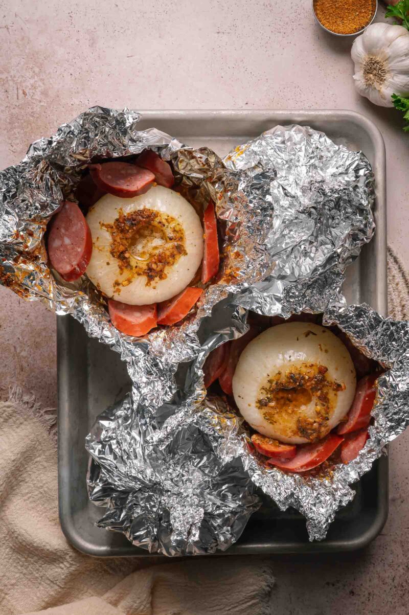 Two foil packets on a baking tray, each containing sliced sausage and a whole onion with seasoning on top.