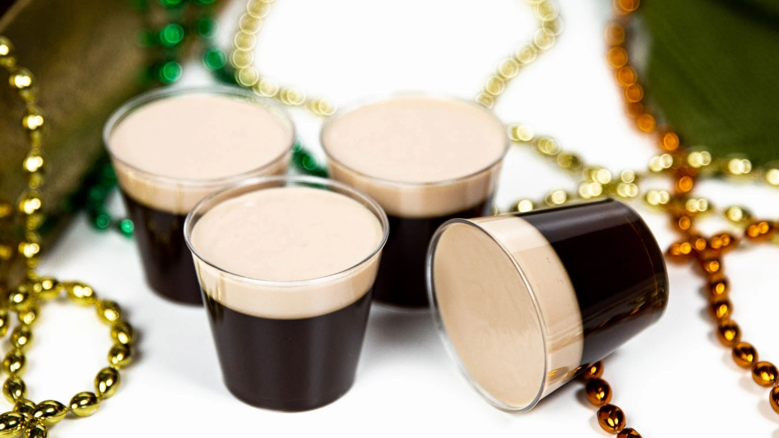 Four small clear plastic cups filled with a dark layered dessert topped with a light cream layer are arranged on a white surface with green, gold, and orange bead necklaces scattered around them.
