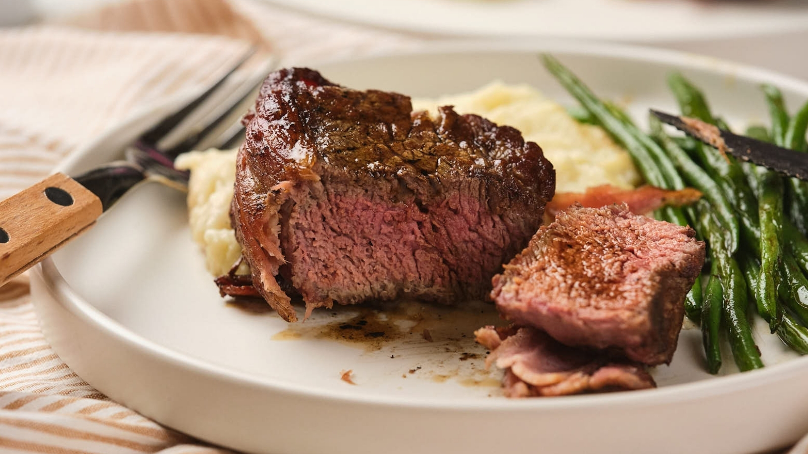A plate with a cooked steak cut into pieces, accompanied by mashed potatoes and green beans.