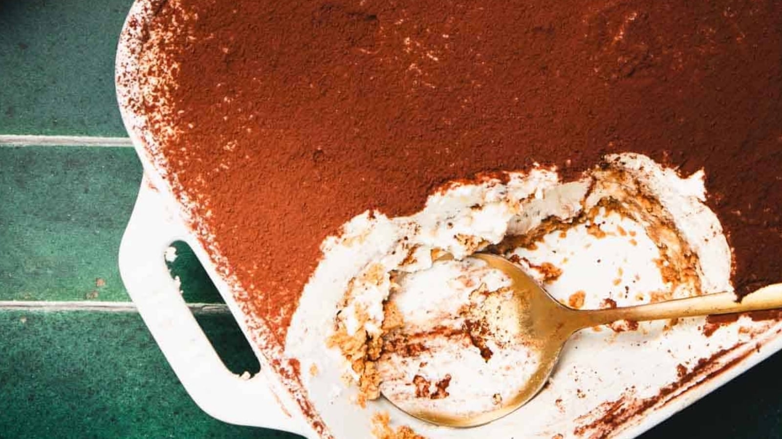 A partially eaten tiramisu in a white baking dish with a gold serving spoon, set on a green tiled surface.