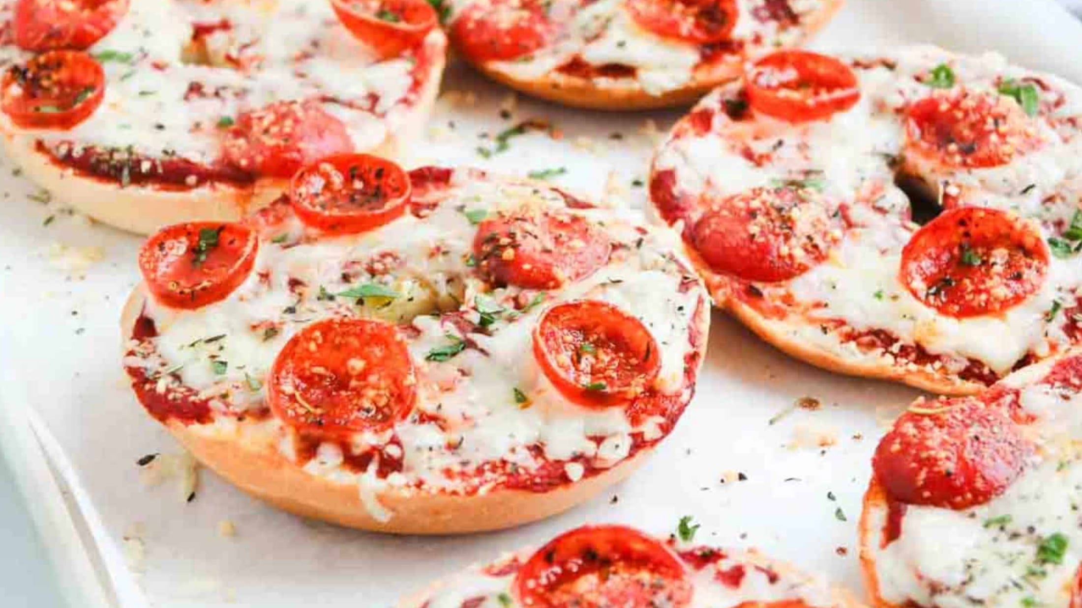 Bagels topped with melted cheese, tomato sauce, and pepperoni, garnished with herbs on a baking tray.