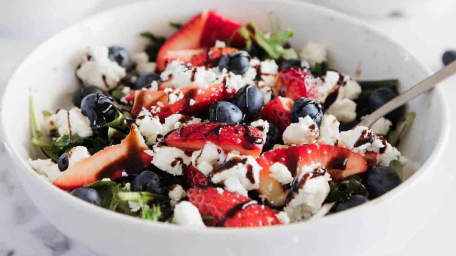 A white bowl filled with a salad of leafy greens, sliced strawberries, blueberries, crumbled white cheese, and a drizzle of dark balsamic glaze, with a metal fork on the right side.