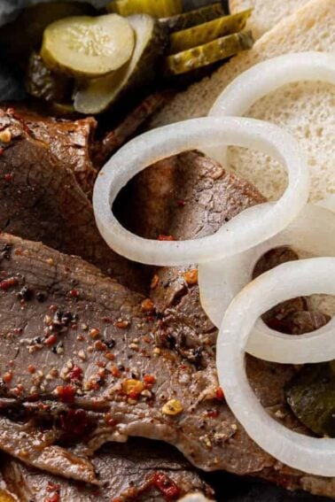 Plate with slices of seasoned brisket, raw onion rings, pickles, and two pieces of white bread on parchment paper.