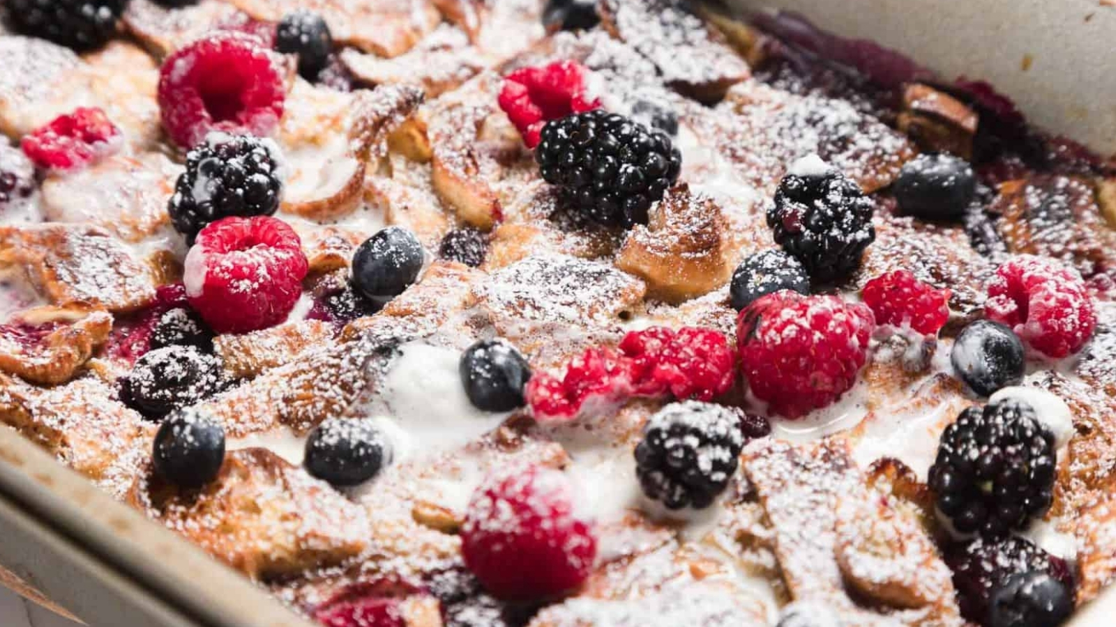 Close-up of a baked dessert topped with powdered sugar, featuring blackberries, raspberries, and blueberries. The dish is in a rectangular baking pan, with visible golden-brown edges and drizzles of cream or sauce.