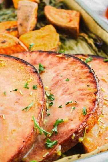 Sliced ham and roasted sweet potatoes on a parchment-lined baking sheet, garnished with herbs.