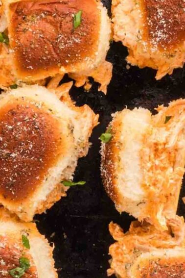 A close-up of several baked sliders filled with shredded chicken and topped with herbs and grated cheese, arranged on a dark surface.