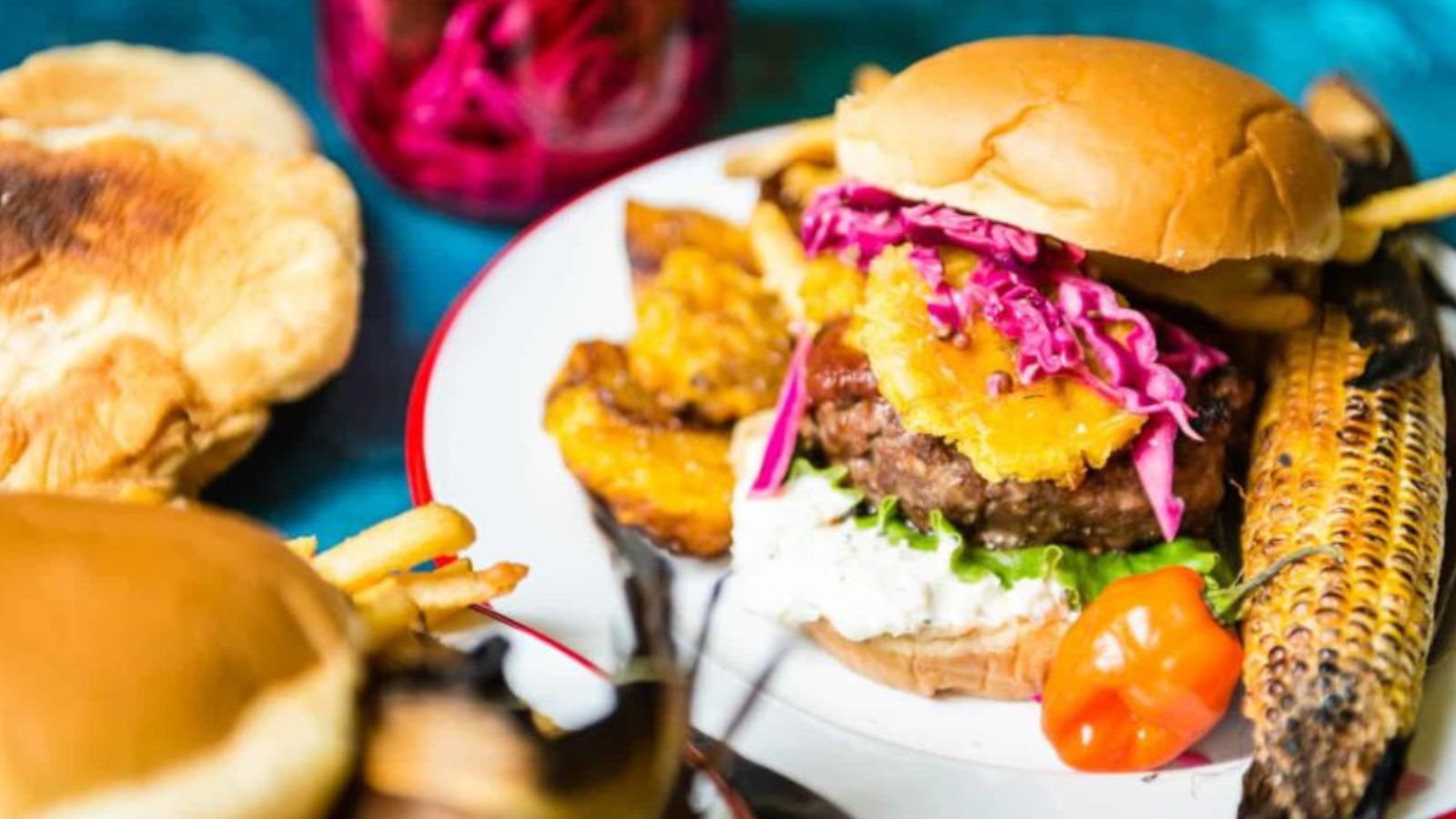 A plated burger with lettuce, slaw, grilled pineapple, and sauce, served with grilled corn, plantains, fries, and a whole pepper; buns and a jar of red slaw are in the background.