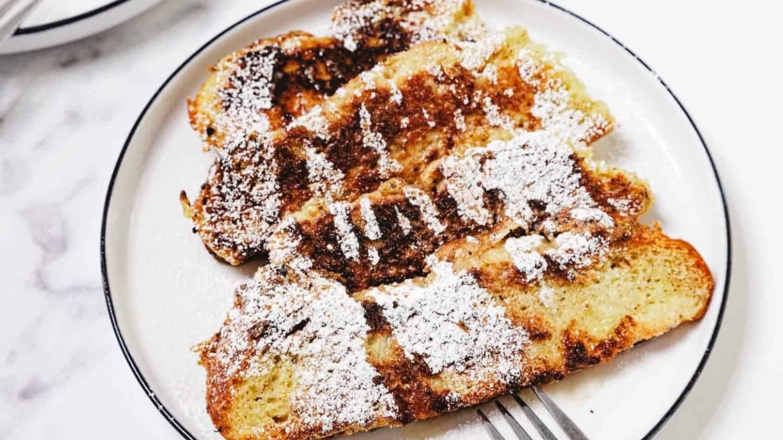 Four slices of French toast topped with powdered sugar on a white plate, with a fork beside them.