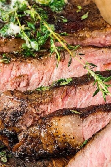 Sliced grilled steak topped with herb butter and garnished with fresh rosemary and thyme on a wooden cutting board.