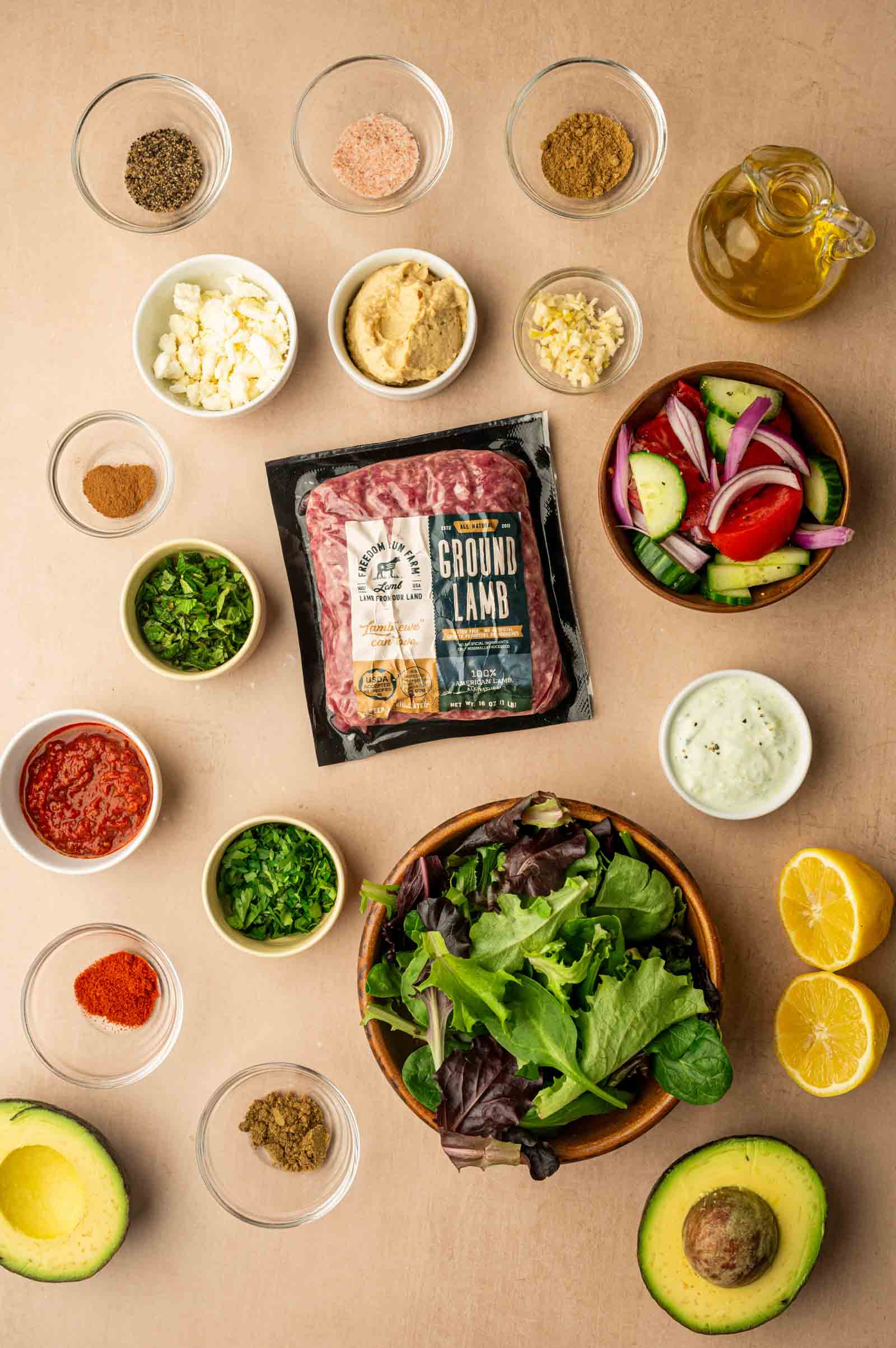 A flat lay of ingredients for a spicy lamb meatball bowl, including ground lamb, fresh vegetables, spices, herbs, sauces, avocado, lemon, and olive oil on a beige surface.
