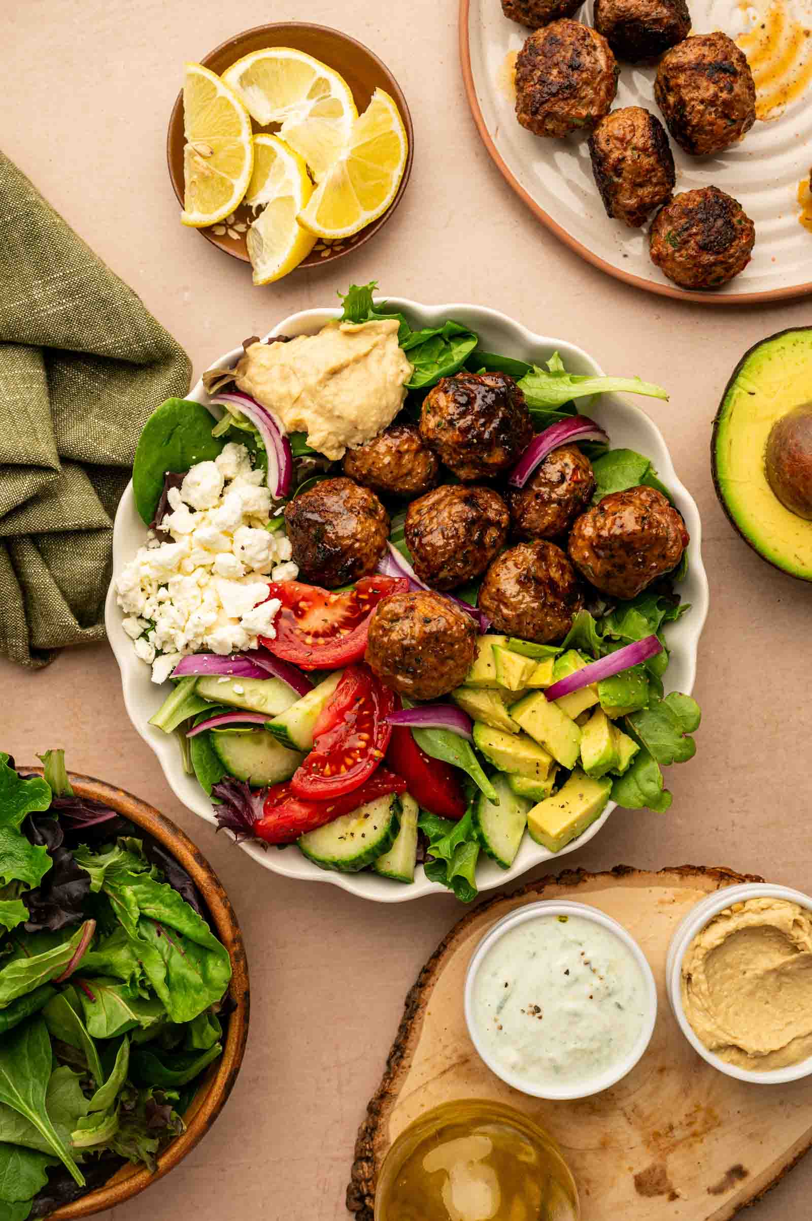 A lamb meatball bowl with vegetables, avocado, feta, hummus, and meatballs is surrounded by plates of meatballs, lemon wedges, avocado, mixed greens, and dips.
