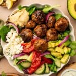 A copycat CAVA spicy lamb meatball bowl with meatballs, mixed greens, tomato, cucumber, avocado, red onion, feta cheese, and a dollop of hummus, surrounded by lemon wedges, avocado, and dressing.