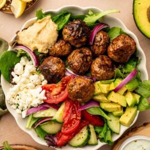 A copycat CAVA spicy lamb meatball bowl with meatballs, mixed greens, tomato, cucumber, avocado, red onion, feta cheese, and a dollop of hummus, surrounded by lemon wedges, avocado, and dressing.