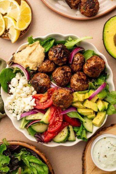 A copycat CAVA spicy lamb meatball bowl with meatballs, mixed greens, tomato, cucumber, avocado, red onion, feta cheese, and a dollop of hummus, surrounded by lemon wedges, avocado, and dressing.