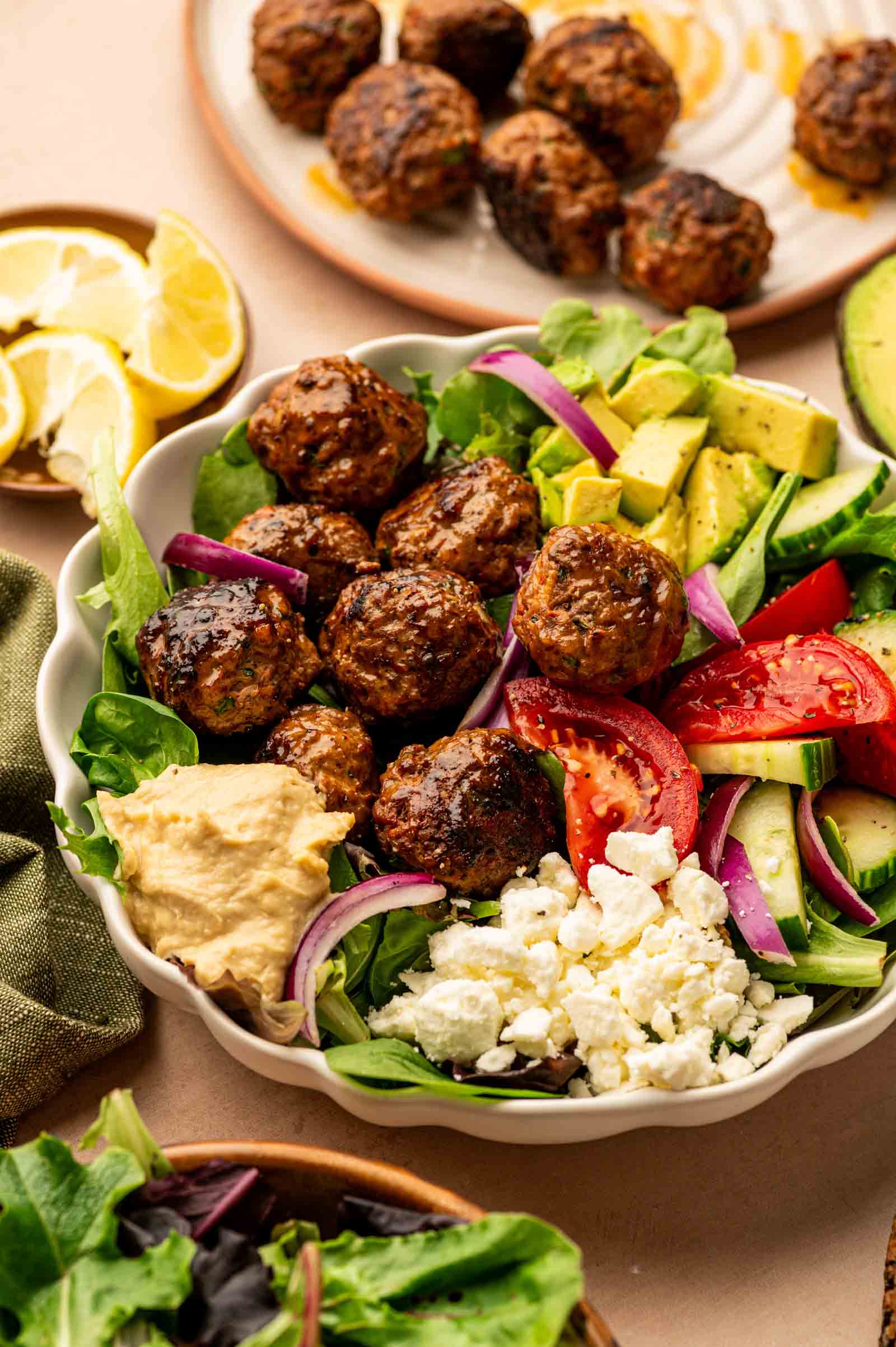 A spicy lamb meatball bowl with meatballs, avocado, tomato, cucumber, red onion, feta cheese, and a scoop of hummus. A plate of meatballs and lemon slices are in the background.