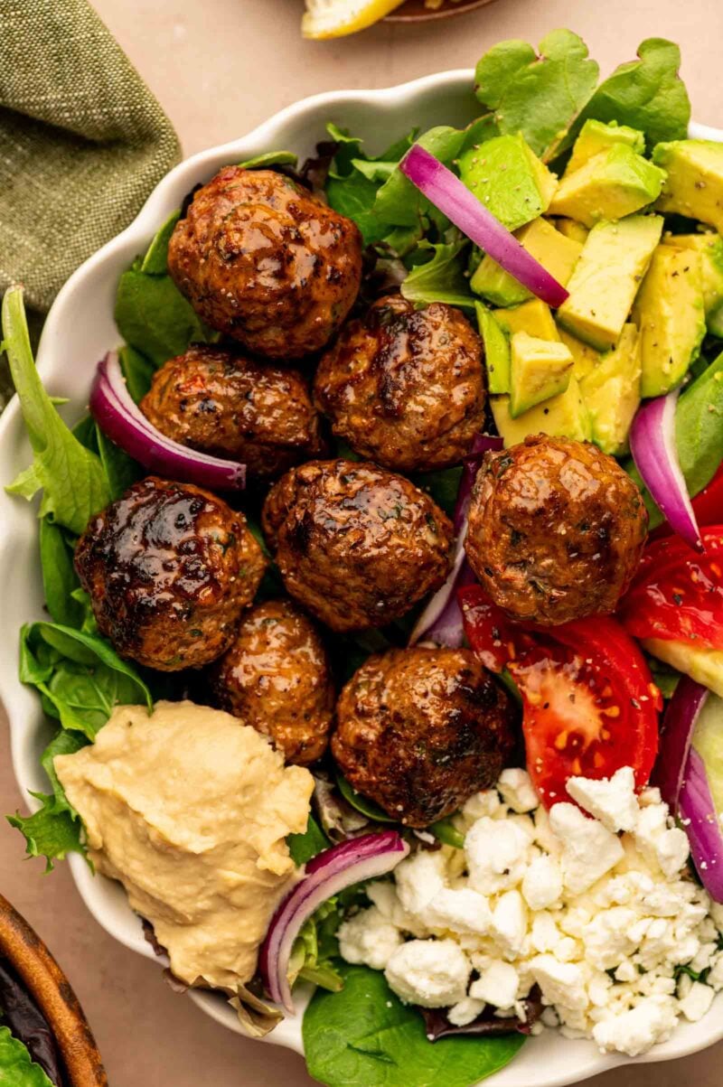 A spicy lamb meatball bowl with meatballs mixed greens, avocado, tomato, red onion, hummus, and crumbled feta cheese.