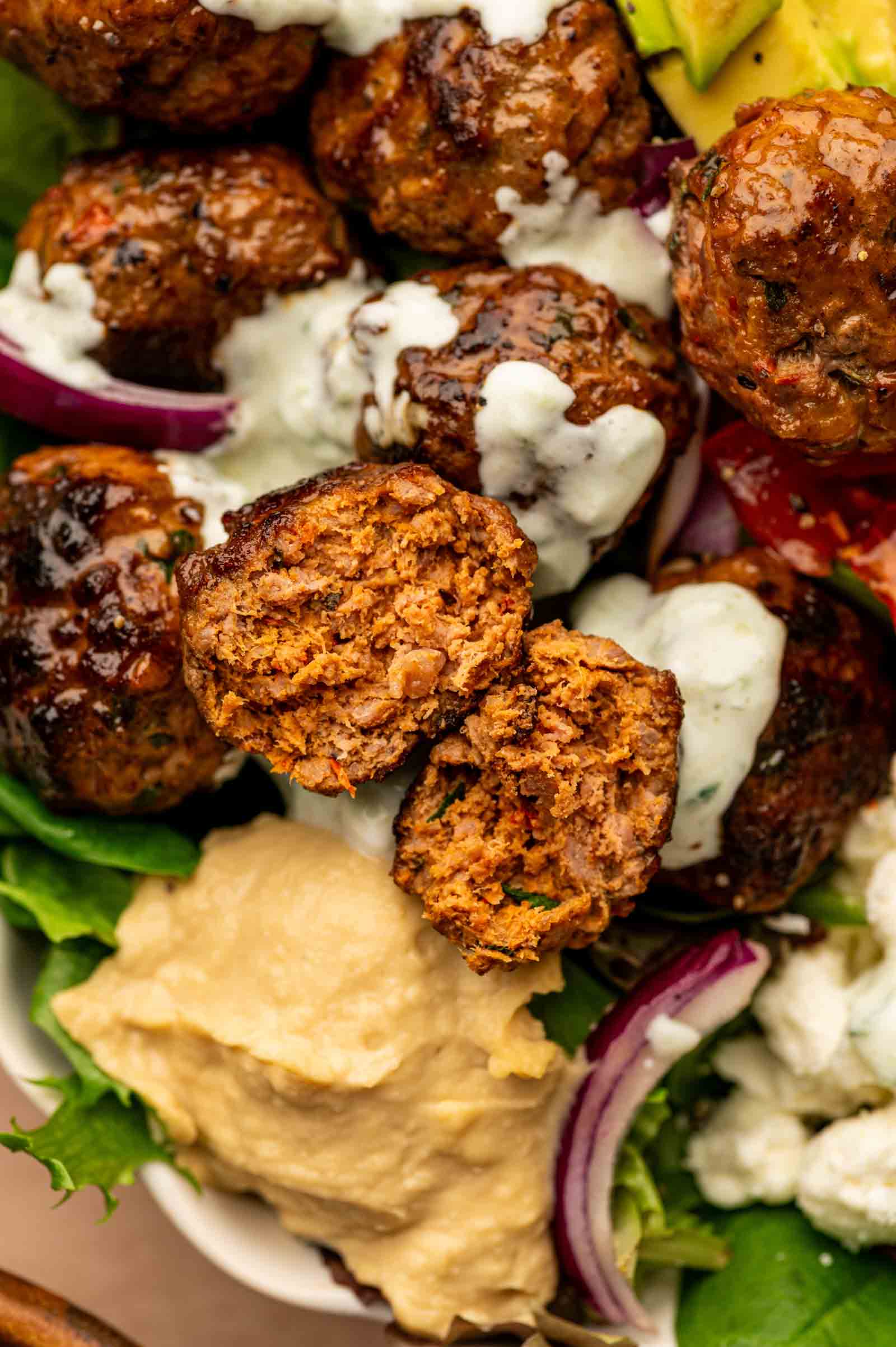A close-up of spicy lamb meatballs with tzatziki sauce, sliced red onions, avocado, lettuce, tomato, a dollop of hummus, and crumbled cheese in a bowl.