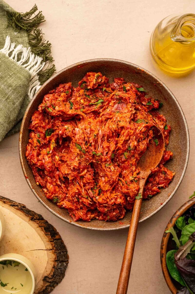 A bowl of seasoned ground lamb with a wooden spoon, surrounded by a green cloth, oil, and salad greens on a beige surface.