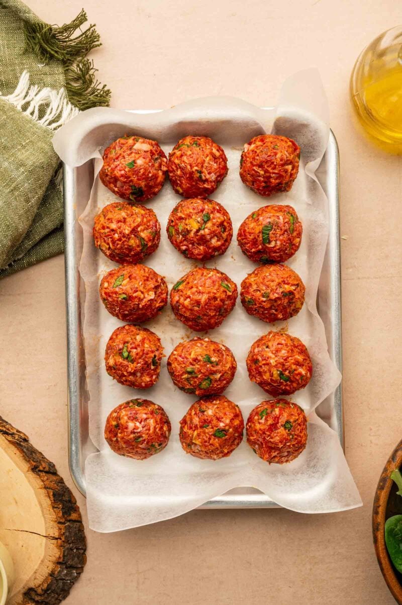 A baking tray lined with parchment paper holding fifteen uncooked meatballs, garnished with herbs, placed on a light surface.