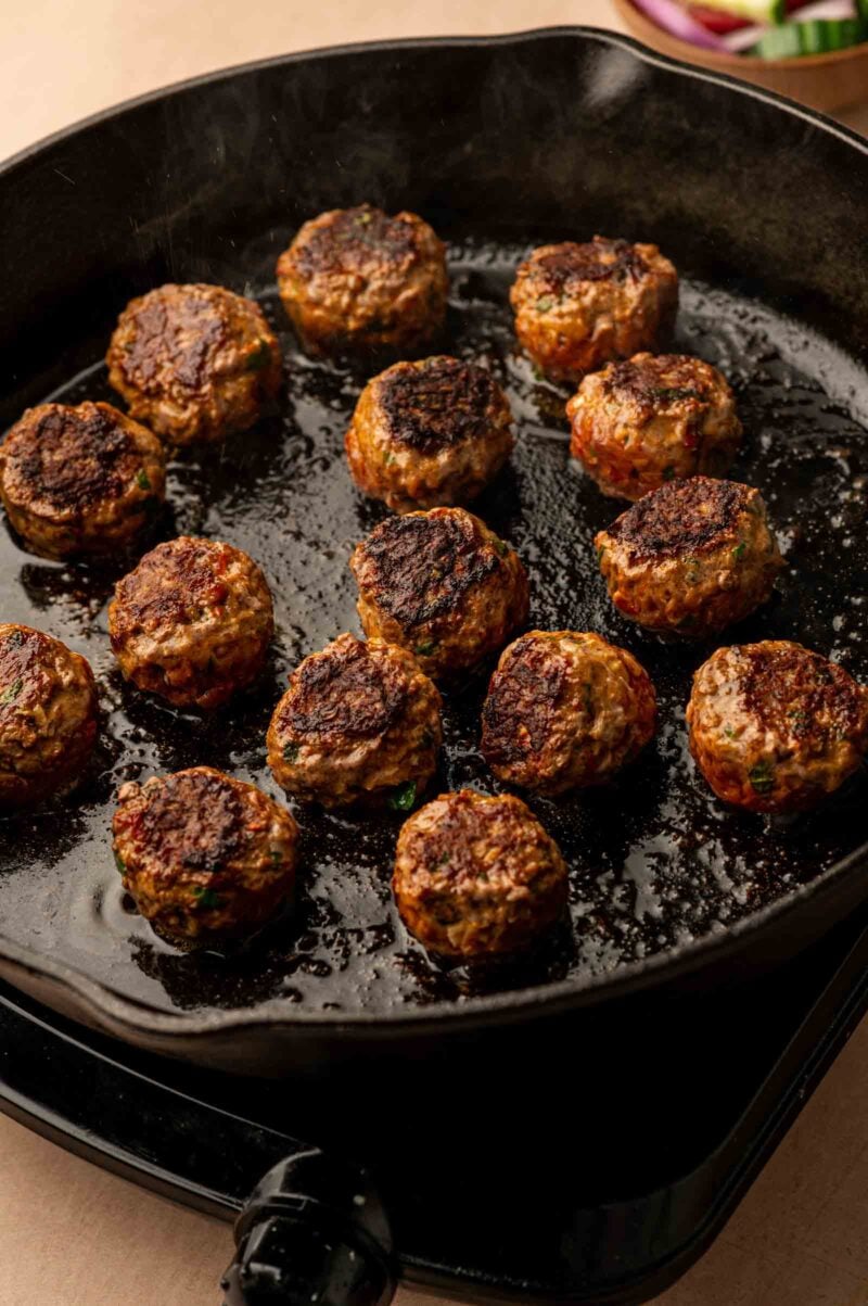Meatballs cooking in a black cast iron skillet, with a browned exterior and visible oil on the surface.