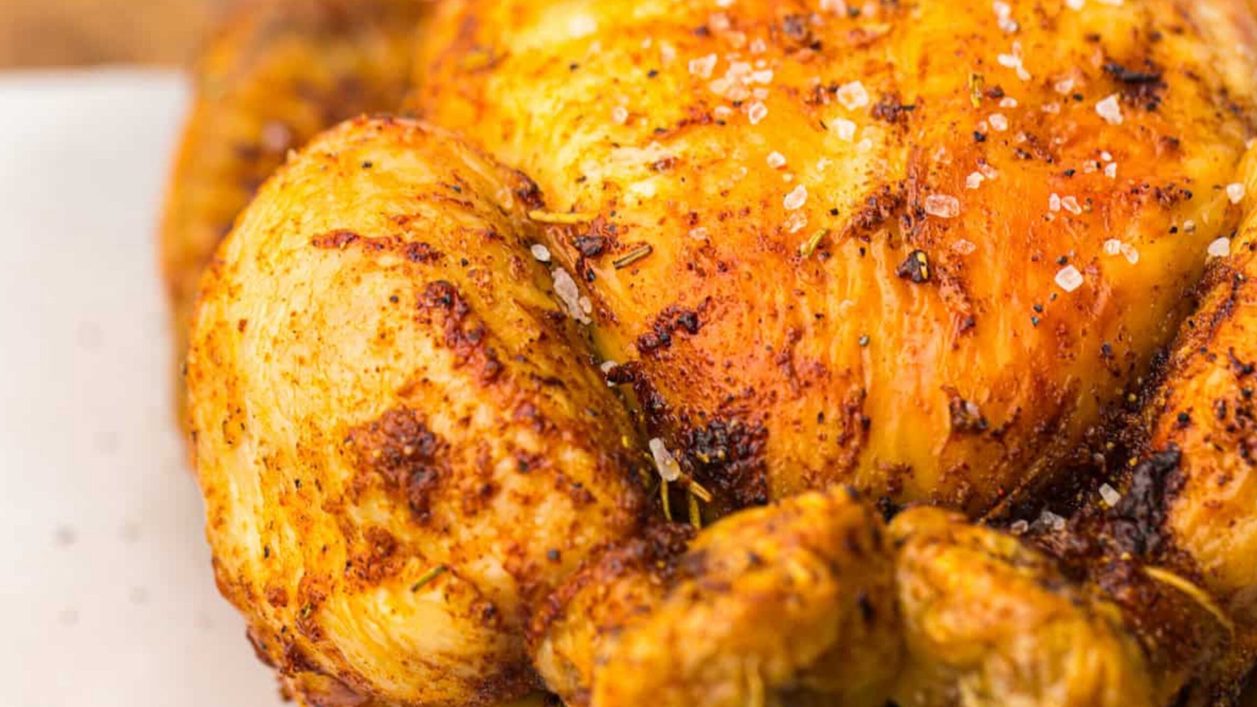 Close-up of a roasted chicken with crispy, golden-brown skin seasoned with herbs and coarse salt, on a wooden cutting board.