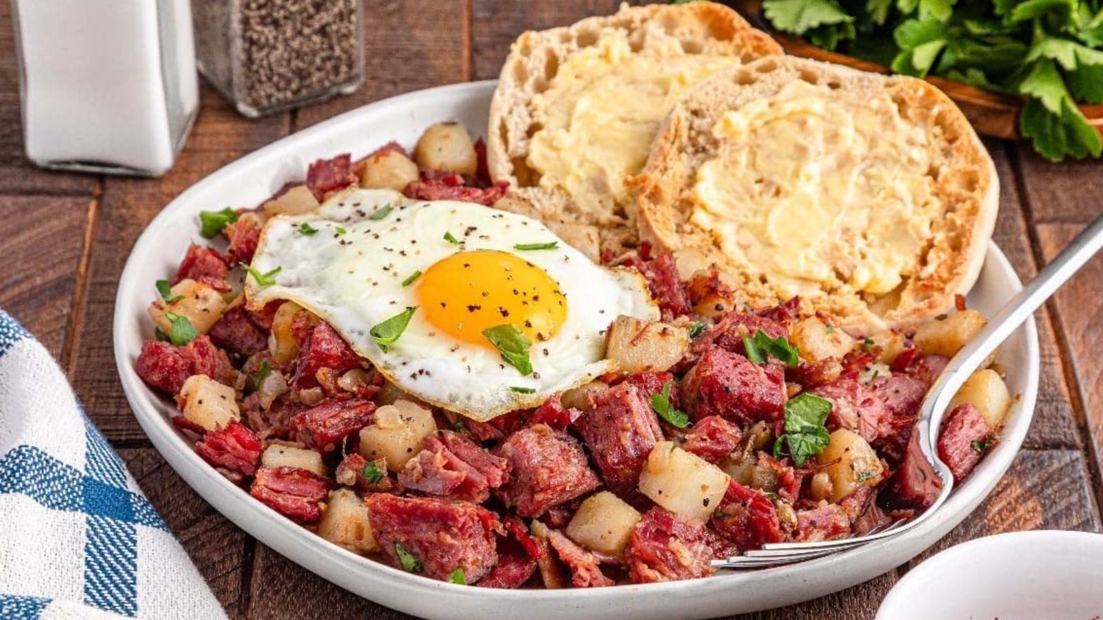 A plate of corned beef hash with potatoes and herbs, topped with a fried egg, served with buttered English muffins on the side.