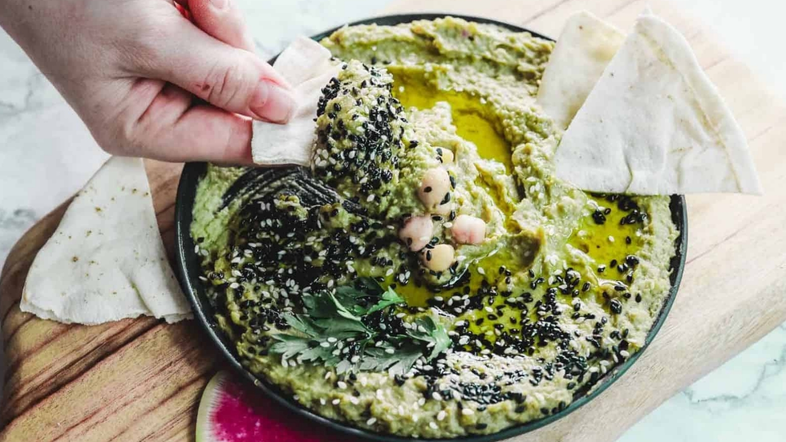 A bowl of green hummus garnished with black sesame seeds, chickpeas, and herbs sits on a wooden board. A hand dips a piece of pita bread into the hummus. Slices of pink and white radish are visible on the board.