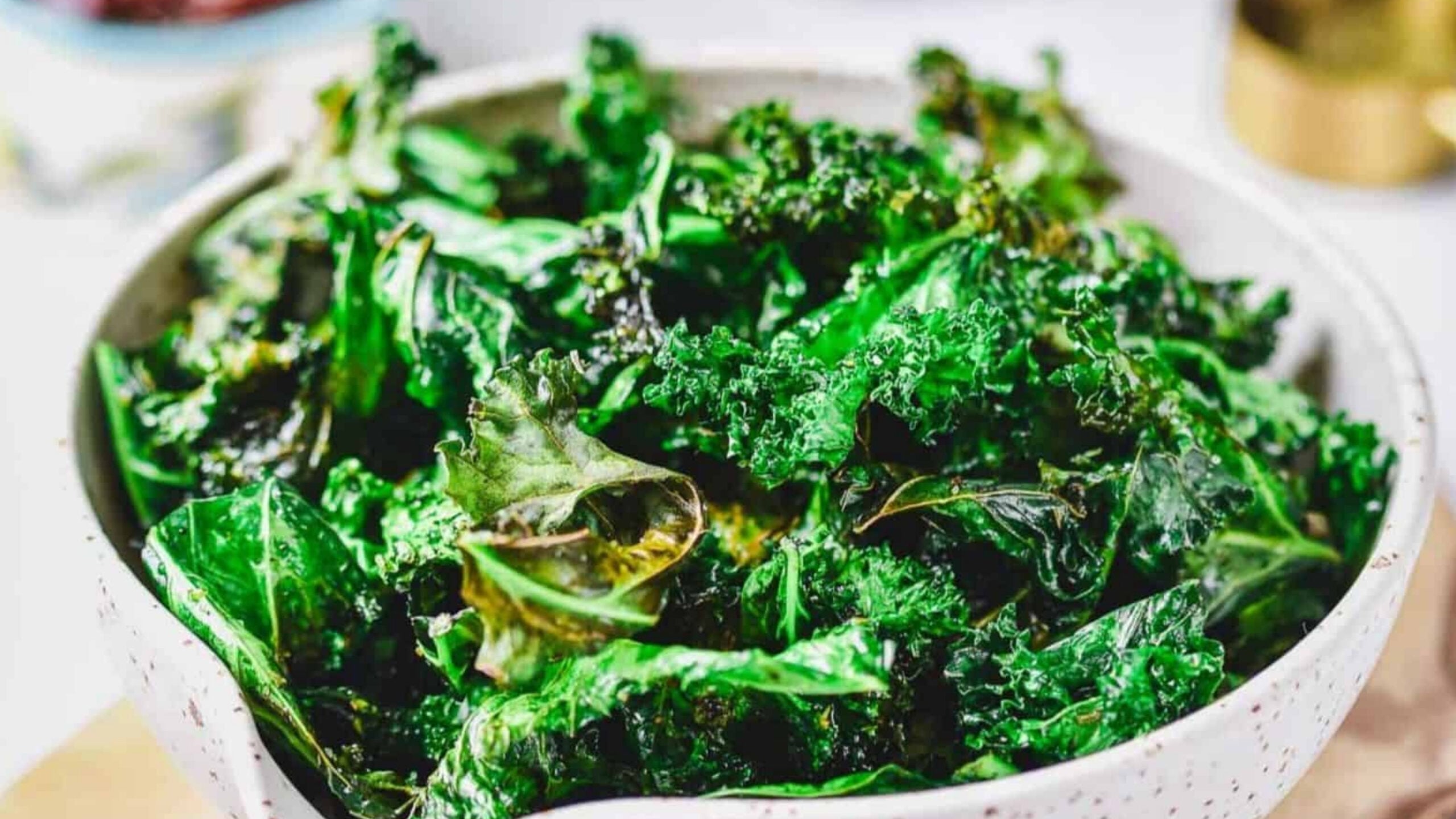 A white speckled bowl filled with crispy, dark green kale chips sits on a light surface. The kale chips appear roasted with some edges curled and slightly browned.