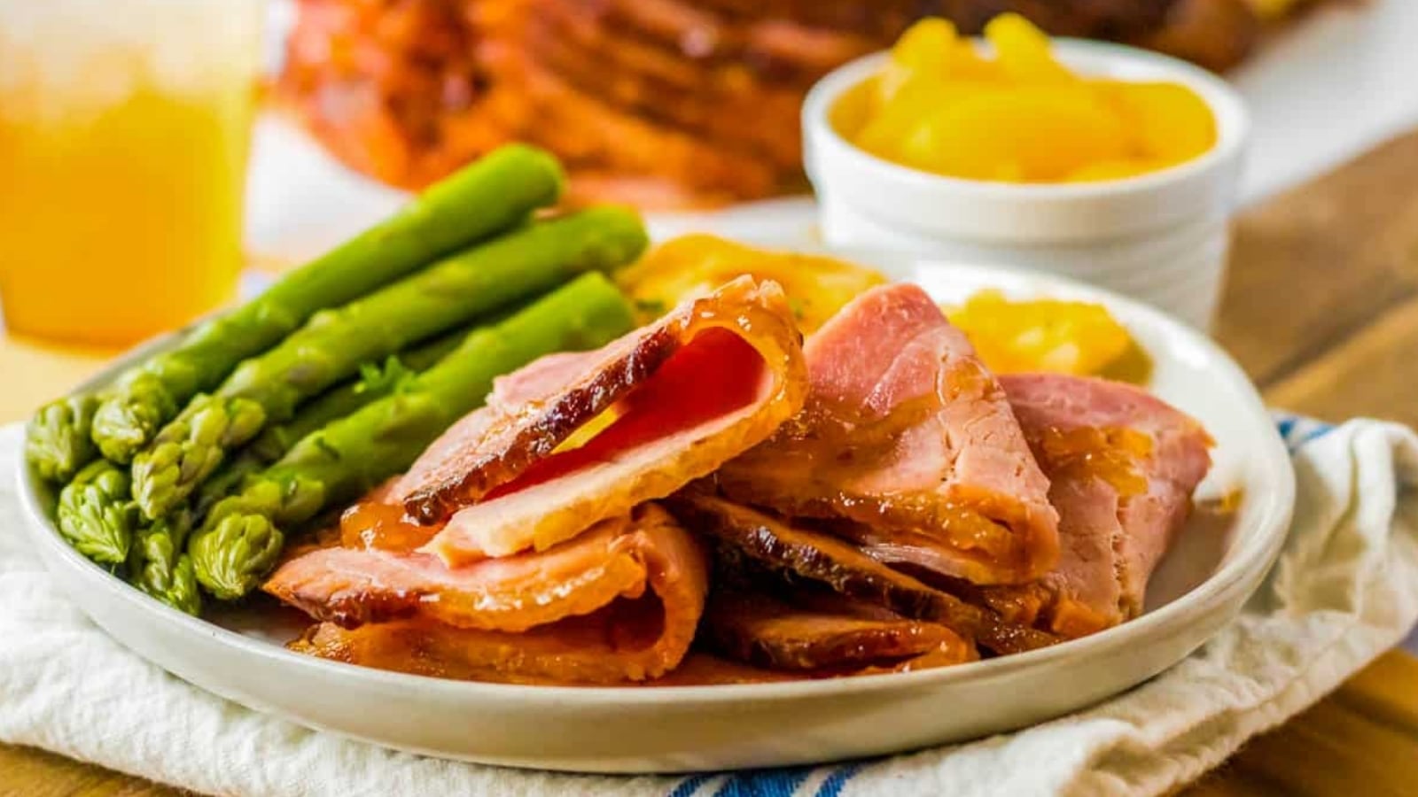 Spiral ham on a plate with asparagus and peaches in the background.