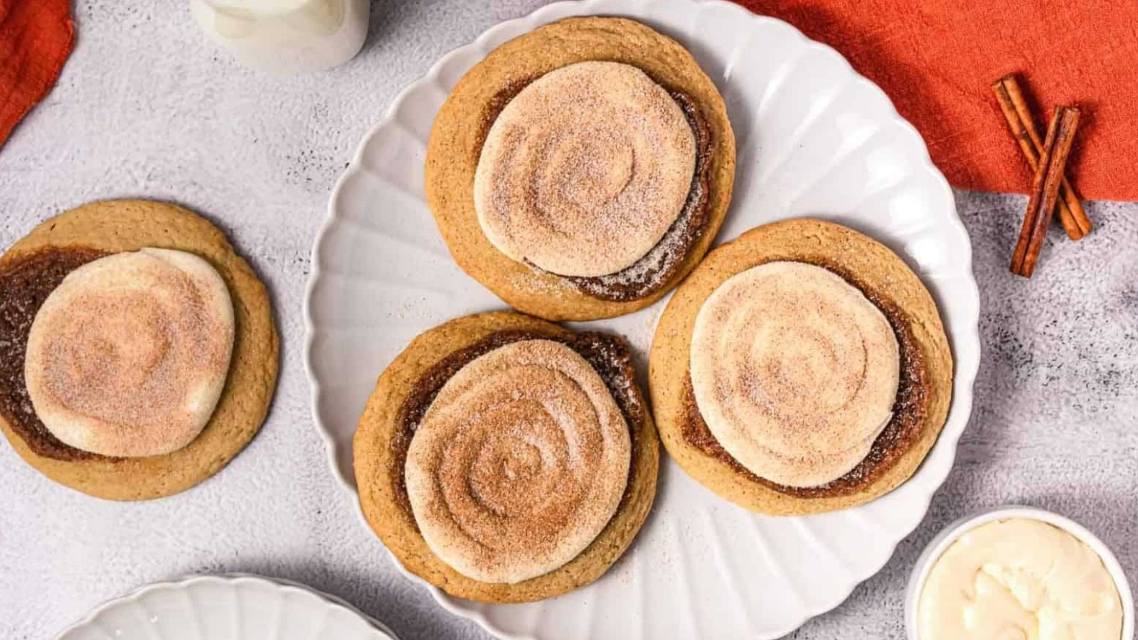 Four cinnamon swirl cookies on a white plate with a glass of milk and cinnamon sticks nearby.