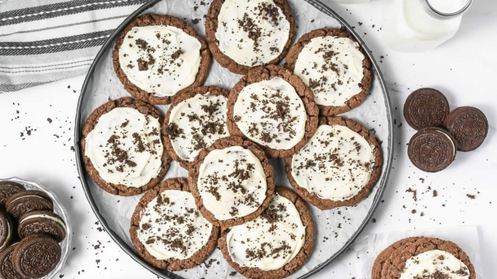 A plate of chocolate cookies with white icing and cookie crumbs, surrounded by sandwich cookies and milk.