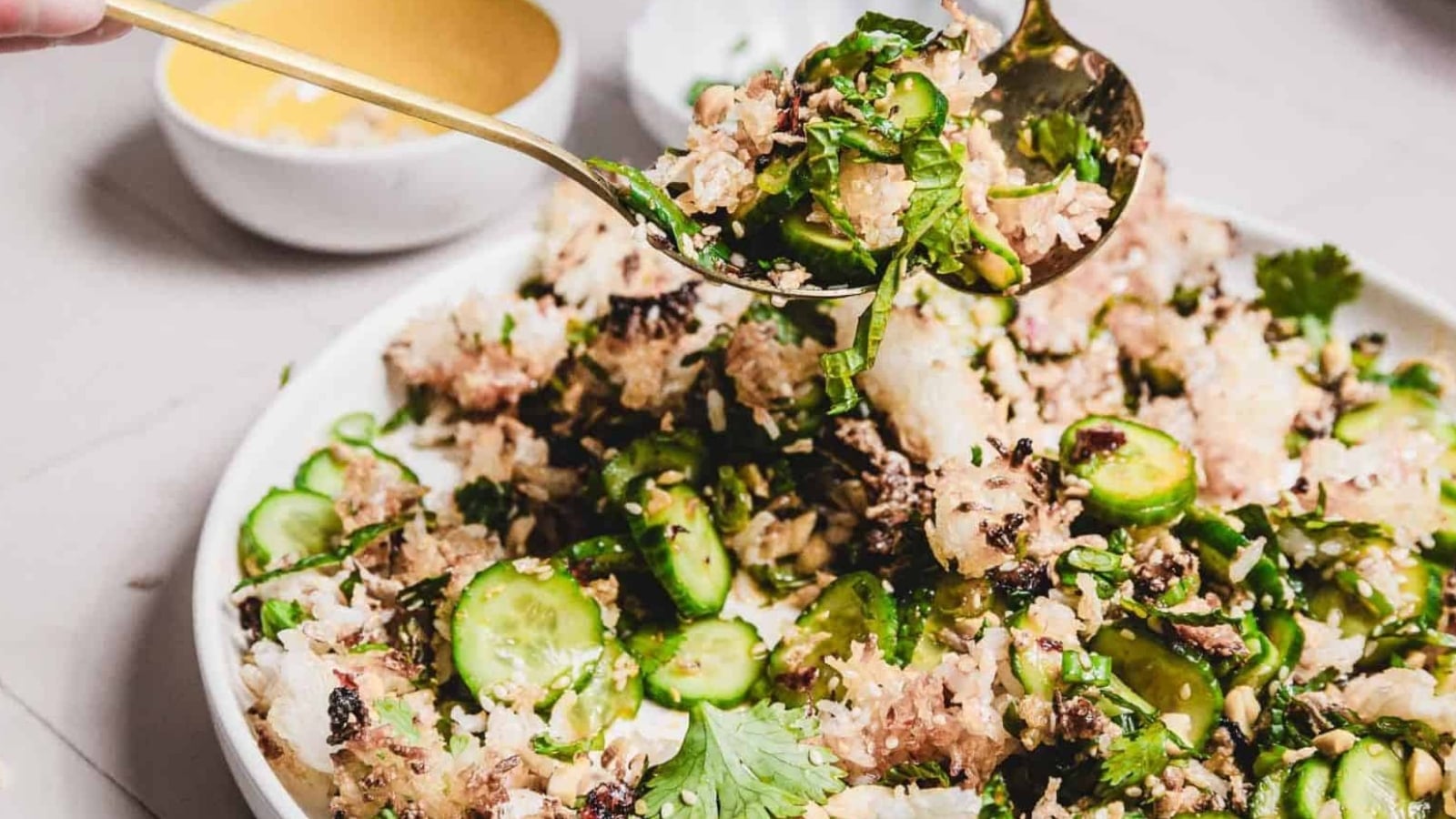 A spoon serving a rice salad with cucumbers, herbs, and crispy toppings from a white plate onto another dish.