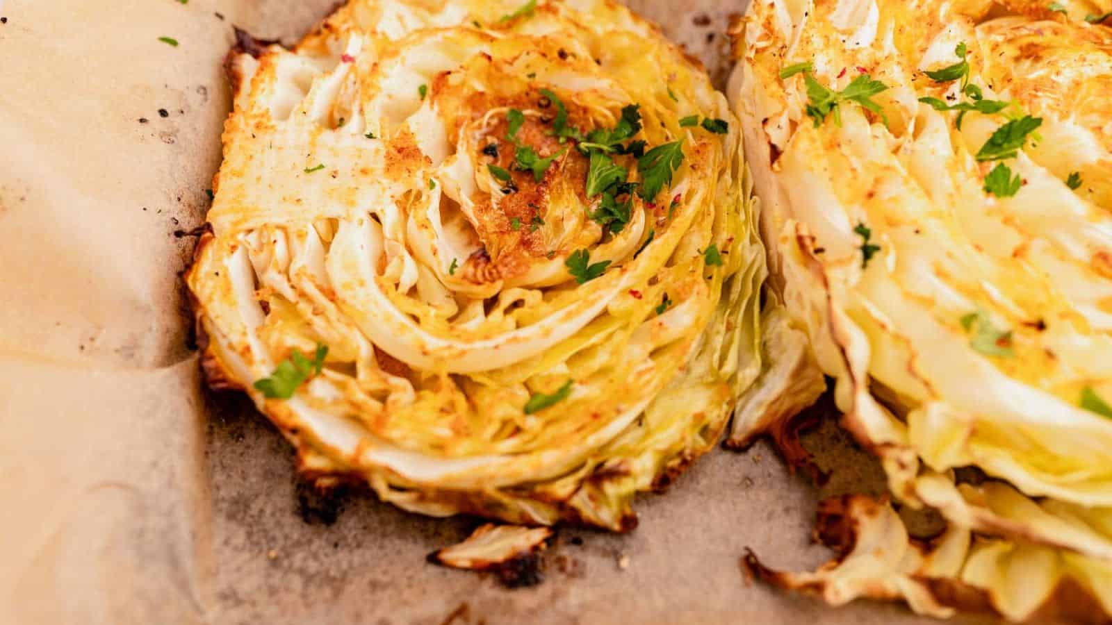 Two thick roasted cabbage slices with browned edges and chopped herbs on parchment paper.