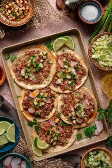 Five dumpling smash taco topped with minced meat, diced vegetables, radishes, and herbs are arranged on a tray with lime wedges, surrounded by various condiments and garnishes.