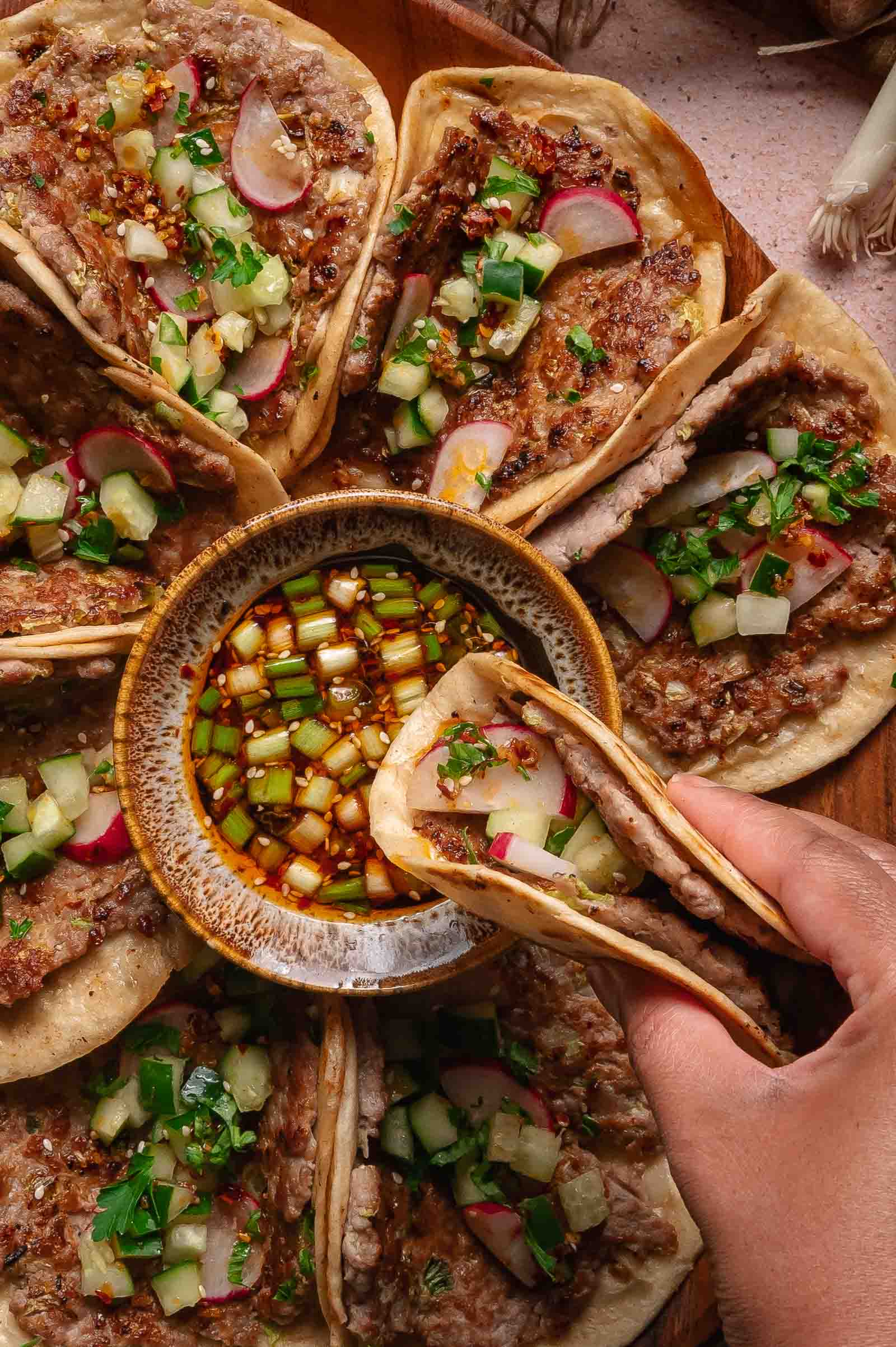 A hand picking up a dumpling smash taco from a plate arranged with several tacos around a bowl of diced vegetables and sauce. The tacos are topped with minced meat and fresh vegetable garnishes.