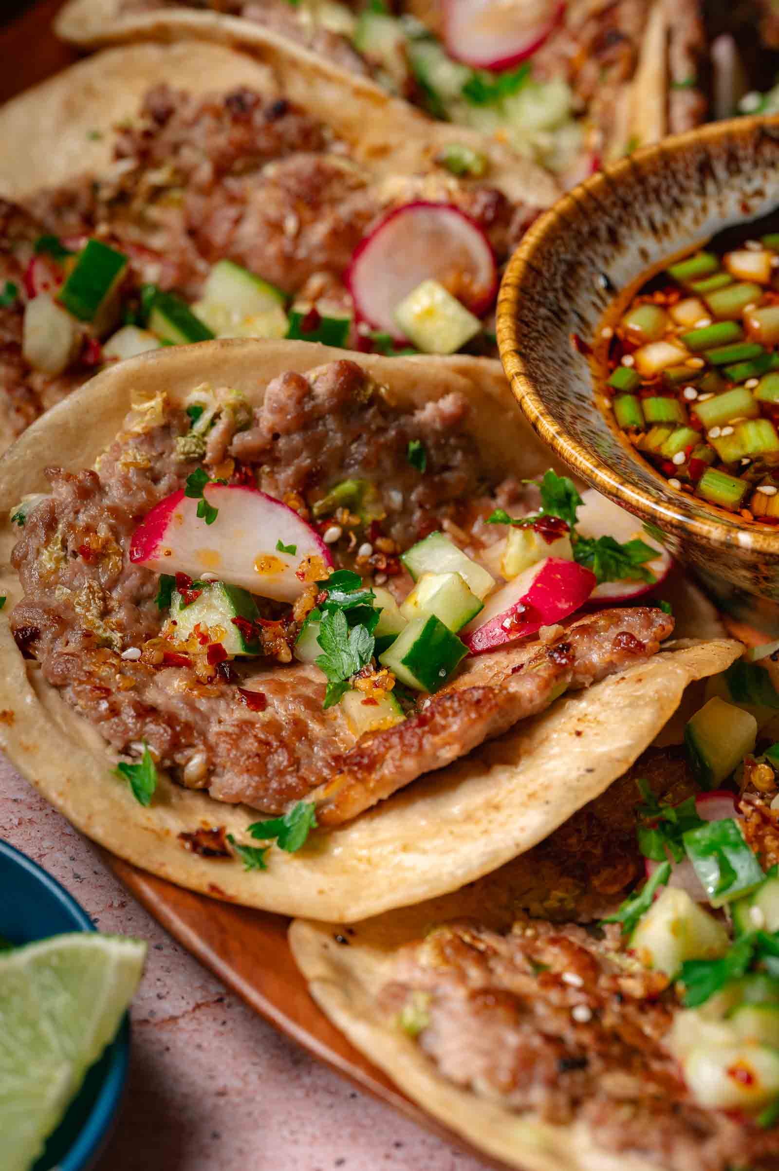 Close-up of dumpling smash tacos topped with chopped radishes, cucumbers, cilantro, and spices, served with a bowl of dipping sauce and a lime wedge on the side.