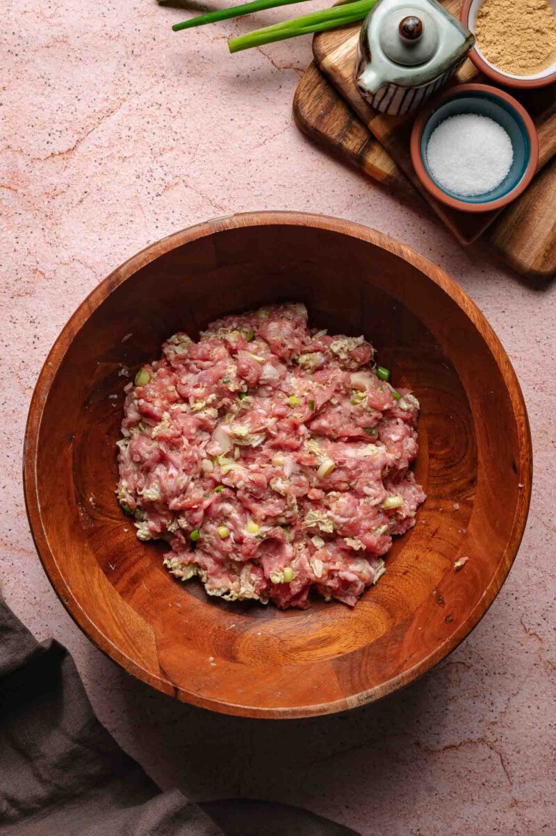 A wooden bowl filled with a dumpkin mixture of raw ground pork, chopped green onions, and seasonings sits on a pink surface next to small dishes of salt, spices, and a soy sauce container.