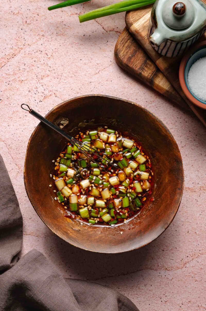 A brown bowl filled with chopped green onions and sauce, with a small whisk inside, sits on a pink countertop next to a teapot, a bowl of salt, and green onion stalks.