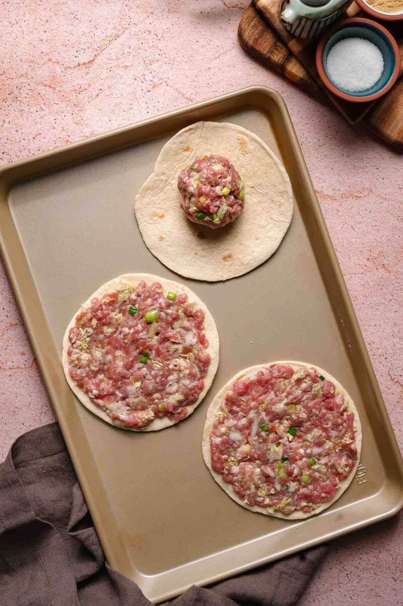 A baking sheet with two raw dumpling smash tacos, an empty tortilla with a ball of filling on top to show pressing the meat evenly. Seasoning bowls and a towel are nearby.