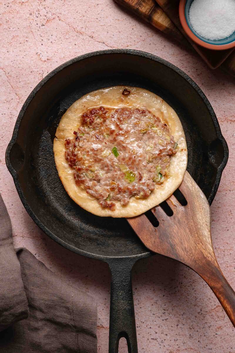 A cooked dumpling smash taco topped with minced meat and green onions sits in a cast iron skillet, with a wooden spatula underneath.