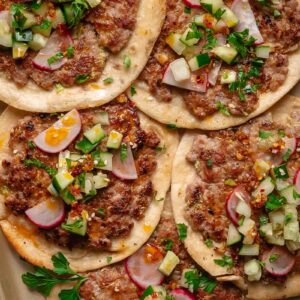 Five dumpling smash taco topped with sliced radishes, chopped cucumbers, onions, herbs, and a sprinkle of chili flakes are arranged on a tray.