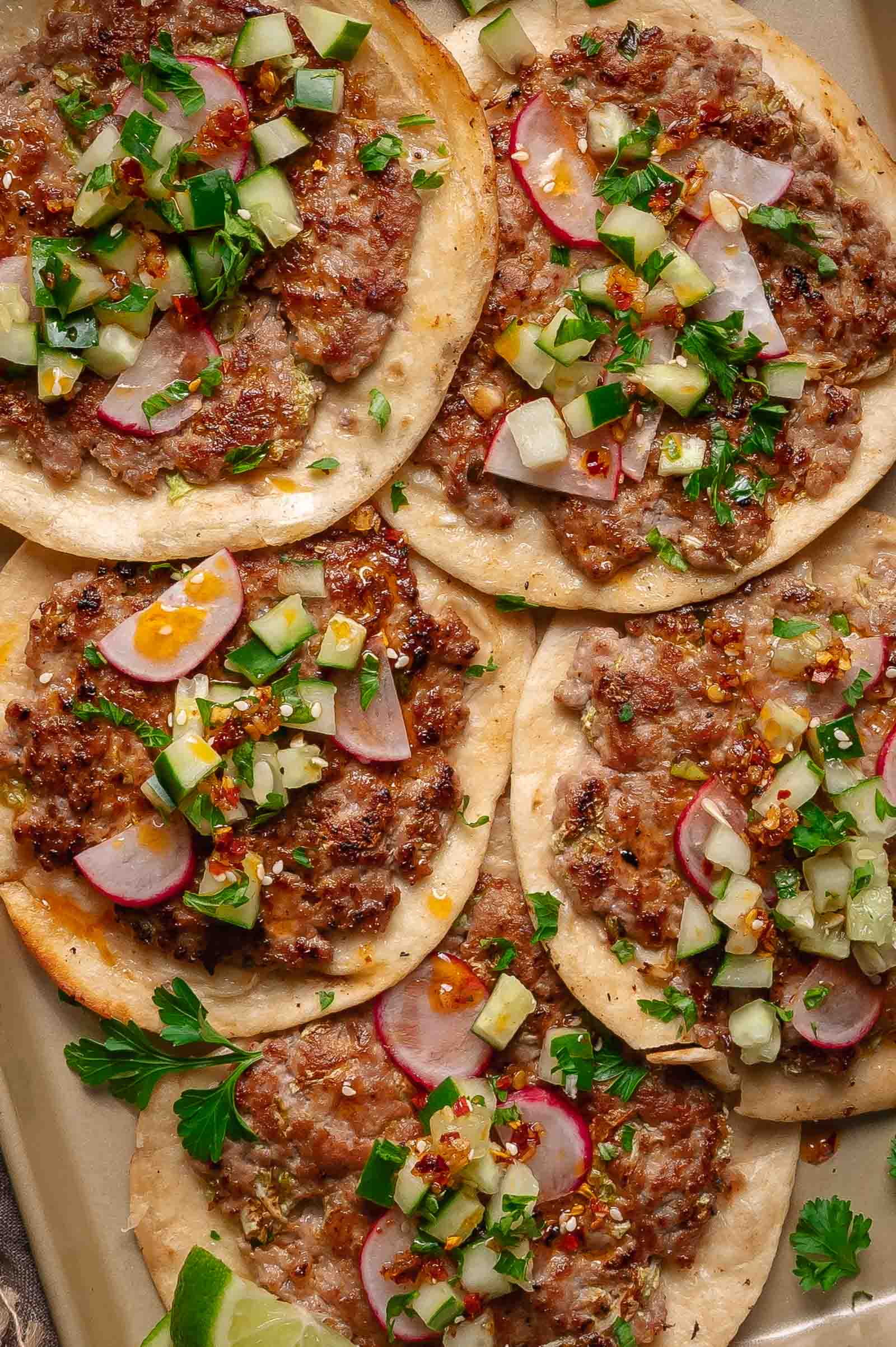 Five dumpling smash taco topped with sliced radishes, chopped cucumbers, onions, herbs, and a sprinkle of chili flakes are arranged on a tray.