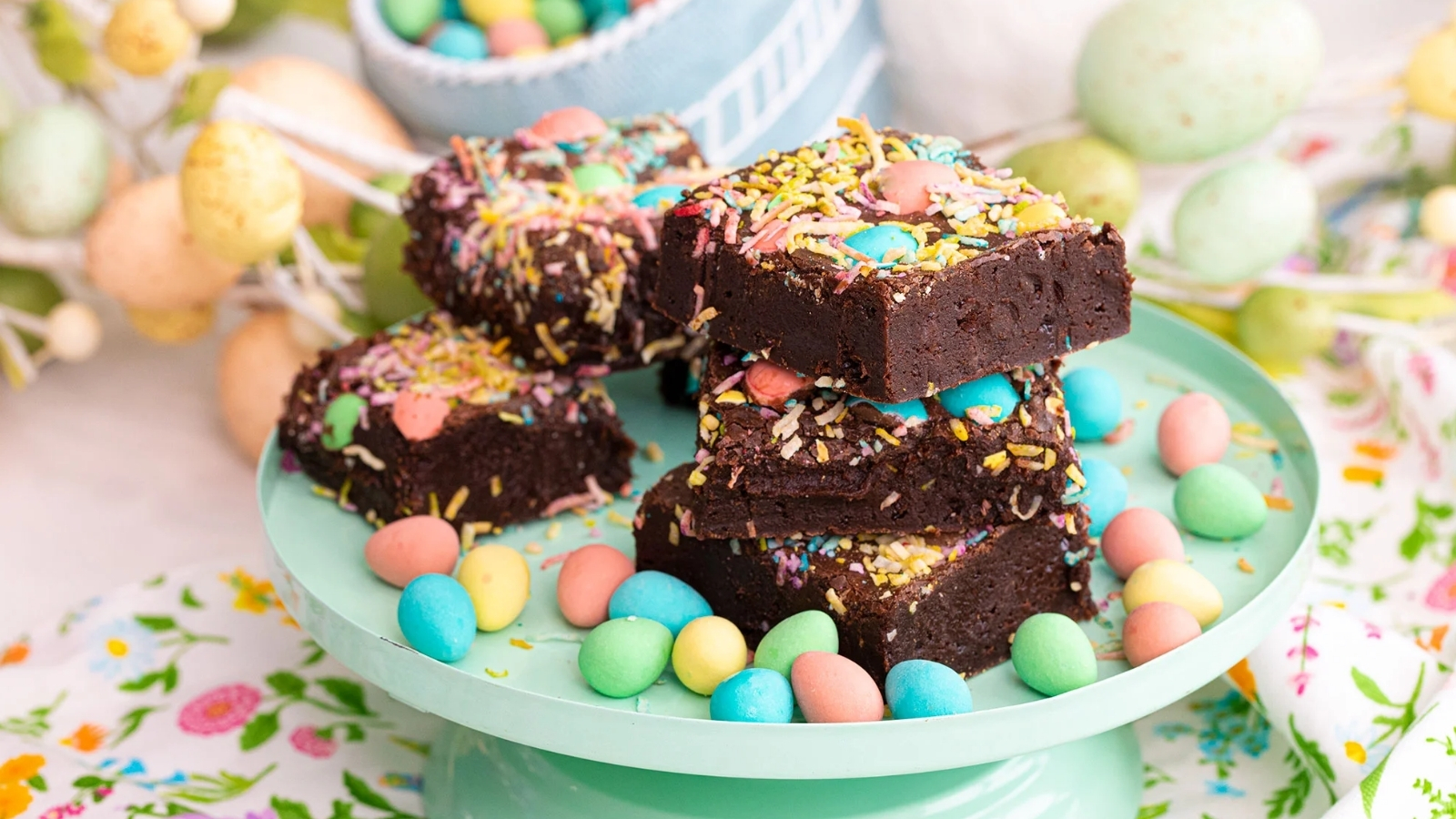 Stacked chocolate brownies topped with colorful sprinkles and candy-coated eggs on a mint green cake stand, with more candy eggs scattered on the plate and a bowl of candies in the background.