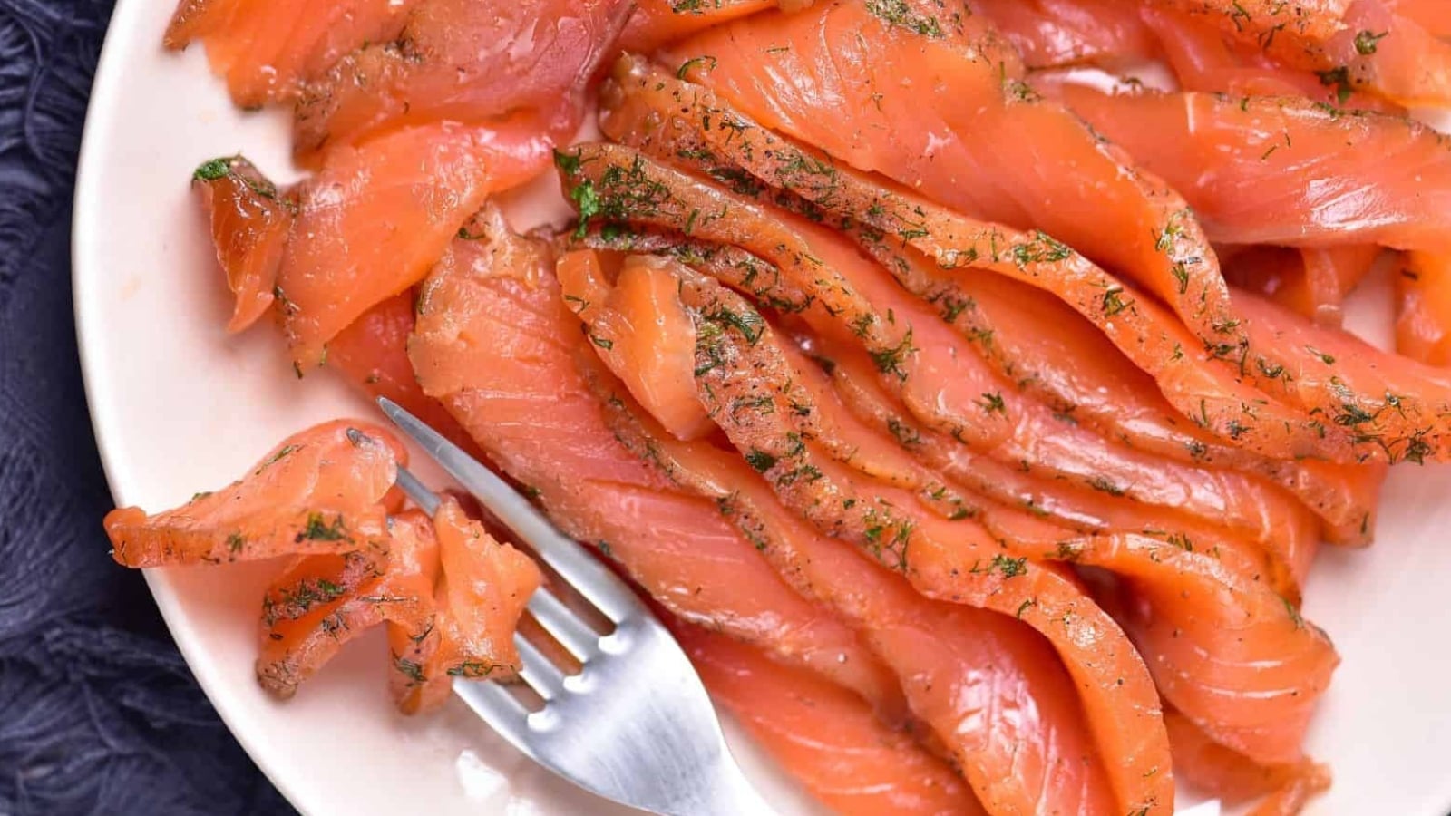 Thin slices of cured salmon with herbs are arranged on a white plate, with a fork holding a piece in the foreground.
