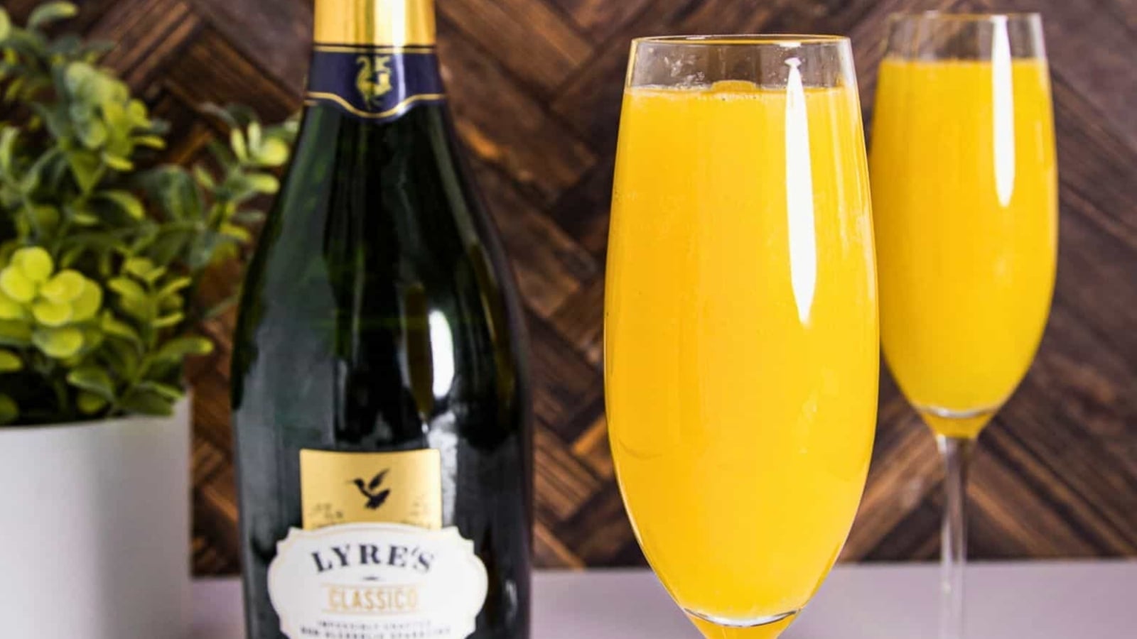 A bottle of Lyre’s sparkling wine next to two champagne flutes filled with orange mimosa; a potted plant is in the background.