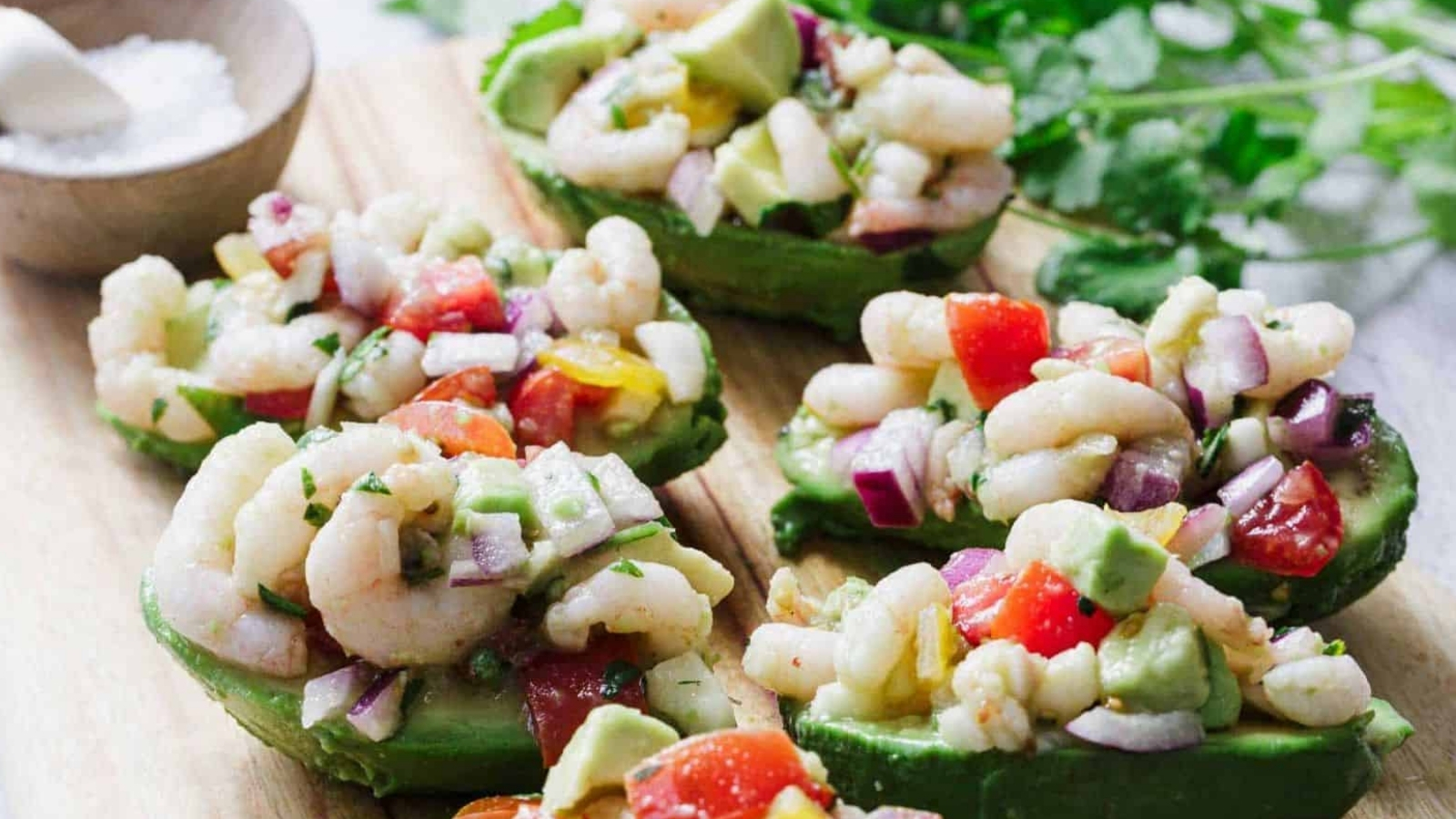 Halved avocados filled with shrimp, diced tomatoes, red onion, avocado chunks, and herbs, served on a wooden board with a bowl of salt in the background.