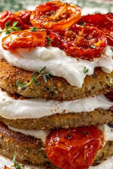 A stack of breaded and fried eggplant slices layered with creamy cheese and topped with roasted cherry tomatoes, garnished with fresh herbs on a decorative plate.